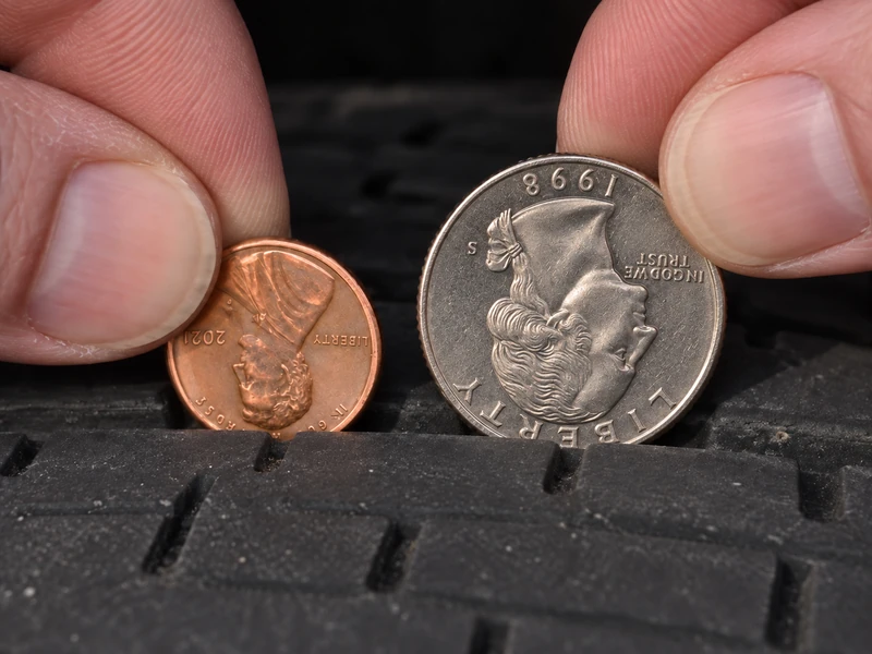 A penny and a quarter held side by side against a tire tread, comparing the 2/32 inch and 4/32 inch tread depth tests